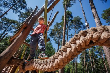 Sakin çocuk, açık mavi gökyüzünün altındaki yüksek ağaçlarla çevrili bir oyun alanındaki halat yapısında güvenle duruyor. Açık havada arkadaşlarıyla güneşli bir günün tadını çıkarıyor.
