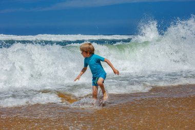 Genç bir çocuk, yaz tatilinde kumsalda güneşli bir günün tadını çıkarırken, neşeyle okyanusa su sıçratır. Etrafı güzel mavi gökyüzü, çarpan dalgalar ve dalgalardan gelen beyaz köpüklerle çevrilidir.