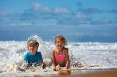 Açık mavi gökyüzü ve köpüklü suyla çevrili güneşli bir plajda oynarken iki çocuk sevinçle okyanus dalgalarında su sıçratıyorlar yaz aktiviteleri için eğlenceli bir ortam yaratıyorlar.