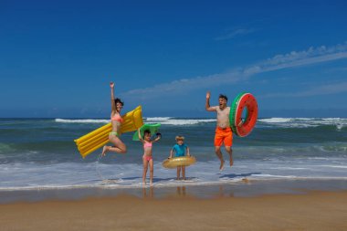 Dört kişilik neşeli bir aile, açık gökyüzü ve kumlu güneşli bir sahilde renkli şişme oyuncaklar tutarken okyanus dalgalarında zıplıyor.