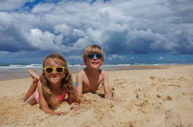 Renkli güneş gözlüğü takan iki çocuk kumsalda kumsalda oynuyorlar. Hafif dalgalar ve okyanus kenarında güzel bir yaz manzarası yaratan dramatik bulutlu bir gökyüzü.