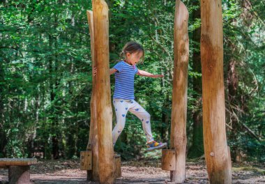 Genç bir çocuk, bereketli yeşil orman oyun parkında ahşap direklerde ustalıkla denge kuruyor, yaz tatilinde ağaçlarla çevrili doğal bir ortamda oyun ve maceranın tadını çıkarıyor.