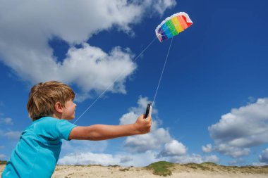 Genç bir çocuk kumsalda neşeli bir gökkuşağı uçurtması uçuruyor, güneşli bir günün tadını çıkarıyor. Arka planda kabarık bulutlar ve saçlarında okyanus esintisi var.