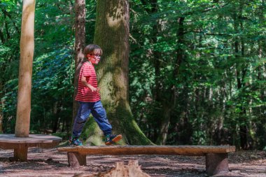 Kırmızı çizgili gömlekli küçük bir çocuk dingin bir orman ortamında yemyeşil ağaçlarla çevrili ahşap bir denge direğinde güvenle yürüyor.