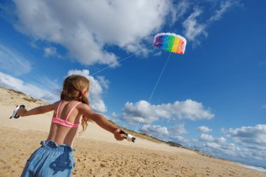 Genç bir kız neşeli bir şekilde güneşli bir sahilde gökkuşağı uçurtması uçuruyor, sıcak havanın ve güzel mavi gökyüzünün tadını çıkarıyor.