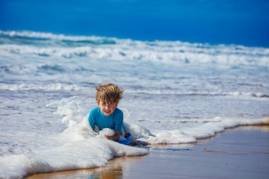 Genç bir çocuk, kumlu sahilde otururken, arka planda köpüklü su ve parlak mavi gökyüzü ile neşeyle oynuyor.