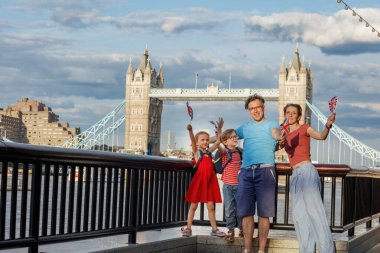 Thames nehrinin kıyısında neşeli bir aile duruyor. Arka planda Tower Bridge olan küçük İngiliz bayrakları sallıyorlar. Parçalı bulutlu bir gökyüzünün altında.