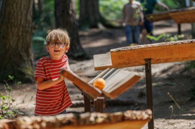 Gözlüklü bir çocuk ormandaki ahşap yapılarla etkileşimden zevk alırken, doğa ile çevrili diğer çocuklar açık hava aktiviteleri ve keşiflere katılıyorlar.