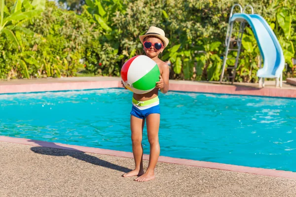 Niño en sombrero sosteniendo la pelota de pie cerca de la piscina —  Fotos de Stock