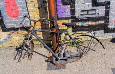Eski bisiklet parçaları güneşli şehir sokağındaki renkli grafiti duvarının önünde paslı direğe kilitlenmiş..