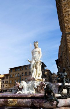 Canlı bir İtalyan şehir meydanındaki süslü bir çeşmenin üzerinde Neptün kulelerinin heykeli. Su, sıcak taş binalar ve parlak mavi gökyüzü tarafından çerçevelenmiş heykel atların ve figürlerin etrafına fışkırır..