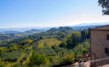 San Gimignano 'nun panoramik manzarası. Terracotta çatıları, tarihi bir kilise ve açık mavi gökyüzü altında yuvarlanan Toskana tepeleri..