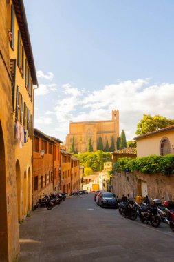Siena 'da San Domenico Bazilikası' na doğru giden büyüleyici bir cadde. Etrafı sıcak tonlu binalar, yeşillikler ve parlak gökyüzünün altında park etmiş scooterlarla çevrili..