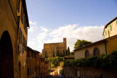 Siena 'da San Domenico Bazilikası' na doğru giden büyüleyici bir cadde. Etrafı sıcak tonlu binalar, yeşillikler ve parlak gökyüzünün altında park etmiş scooterlarla çevrili..