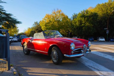 Üstü açık parlak kırmızı bir araba sonbahar güneşiyle yıkanmış bir şehir caddesinde duruyor. Altın ağaçlar arka planda dolaşırken diğer arabalar ve yaya geçidi canlı bir şehir ortamı yaratıyor..