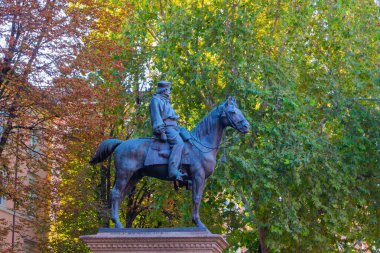 Atlı bir binicinin bronz binicilik heykeli, arka planda renkli sonbahar yaprakları olan bir parkta bir kaidede duruyor..