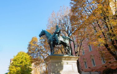 Atlı bir binicinin bronz binicilik heykeli, arka planda renkli sonbahar yaprakları olan bir parkta bir kaidede duruyor..