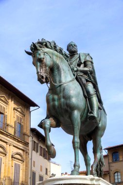 Atlı bir atın bronz heykeli büyük bir İtalyan belediye binasının önünde duruyor. Sütunları oyulmuş, pencereleri kapalı ve güneşli bir meydandaki renkli binaların çerçevelediği ayrıntılı bir cephe..