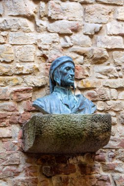 Mavi patinalı bronz bir büst, yıpranmış tuğla bir duvara monte edilmiş sert bir taş kaideye oturur. Heykel, tarihi bir açık hava ortamında asalet ve zamansızlık taşır..