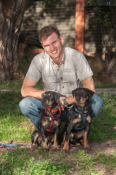 The smiling sitting man in a light shirt keeps two Jagdterriers in full hunting equipment with hand bells — Stock Photo, Image