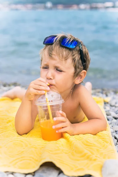 Little boy drinks juice on beach against sea — Stock Photo, Image