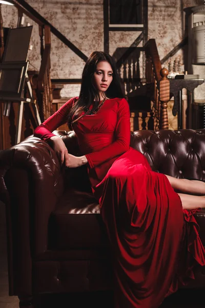 Jovem sensual elegante em vestido vermelho sentado no sofá de couro — Fotografia de Stock