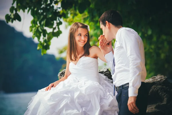 Young bride sits on rock and groom kisses hand — Stock Photo, Image