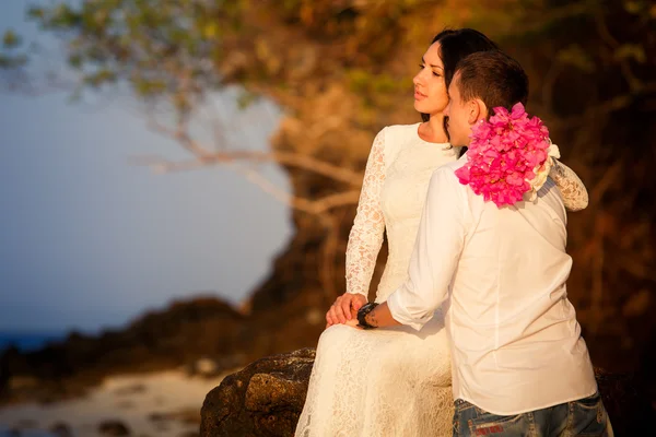 Bride sits on rock groom stands on beach — Stock Photo, Image