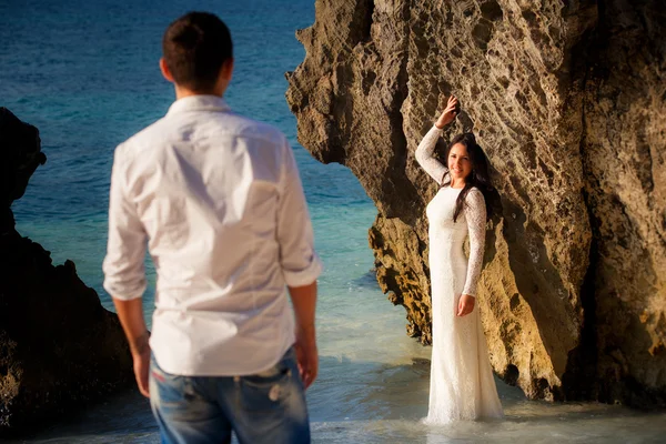 Bride leans against rock on beach groom near — Stock Photo, Image
