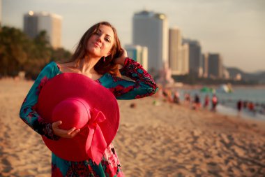 slim girl holds big hat behind back on beach near city sea