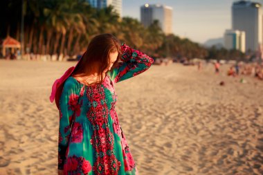 slim girl holds big hat behind back on beach near city sea