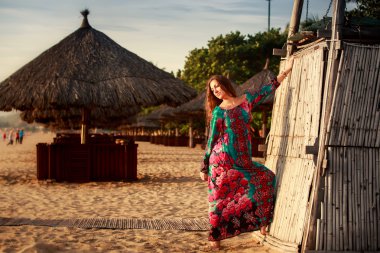 slim girl in long and big red hat leans on reed wall on beach