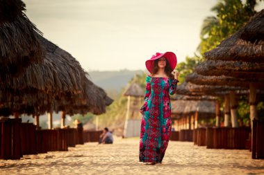slim girl in long and big red hat stands among beach umbrellas