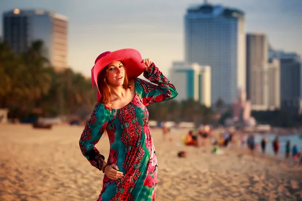 Slim girl in long and big hat on beach against city sea — Stock Photo, Image