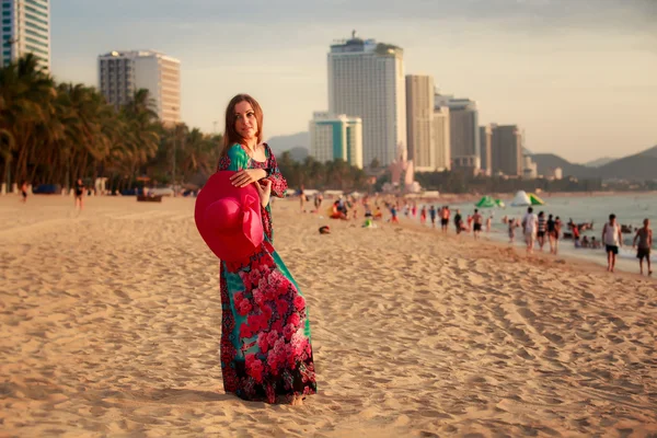 Menina magra em longa detém grande chapéu na praia contra o mar da cidade — Fotografia de Stock