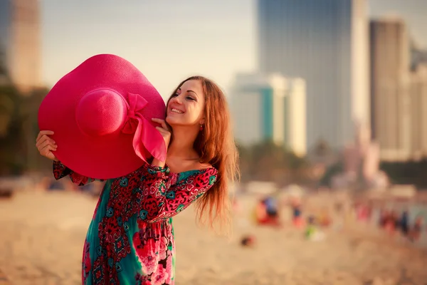 Menina magra em longas mostra grande chapéu na praia contra o mar da cidade — Fotografia de Stock