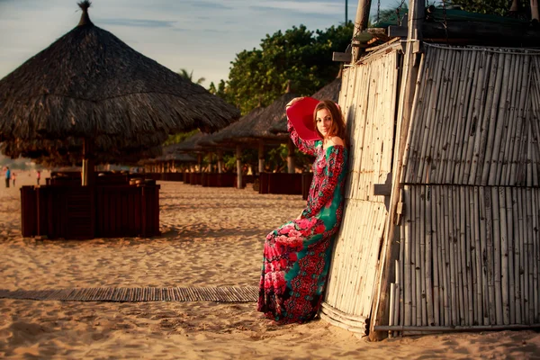 Slim girl in long and big red hat leans on reed wall on beach — Stock fotografie