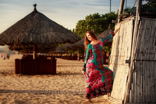 Slim girl in long and big red hat leans on reed wall on beach — Zdjęcie stockowe