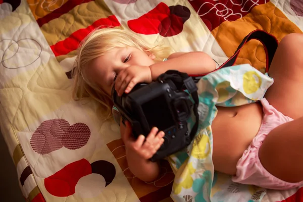 Pequeña chica rubia se encuentra en el sofá examina la cámara fotográfica — Foto de Stock