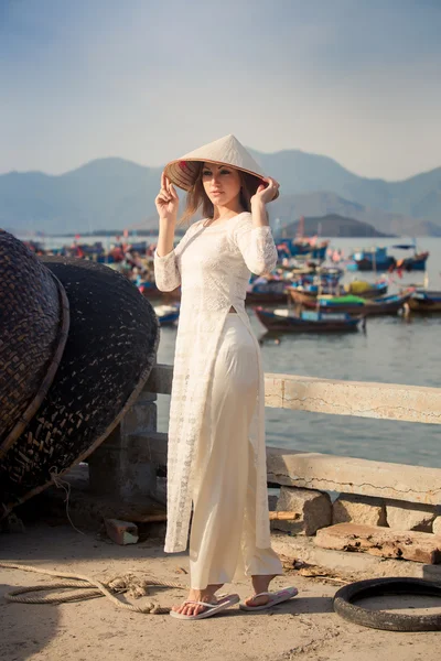 Blonde girl in Vietnamese dress touches hat by barrier — Stock fotografie