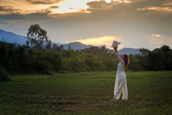 Blonde girl in Vietnamese dress holds hat on field — Stock fotografie