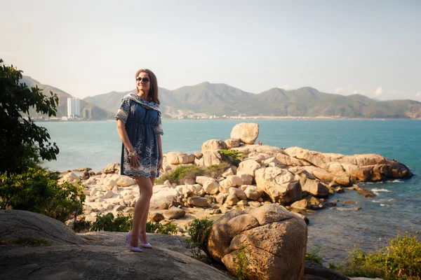 Menina em curto vestido cinza fica em rochas por mar contra a cidade — Fotografia de Stock