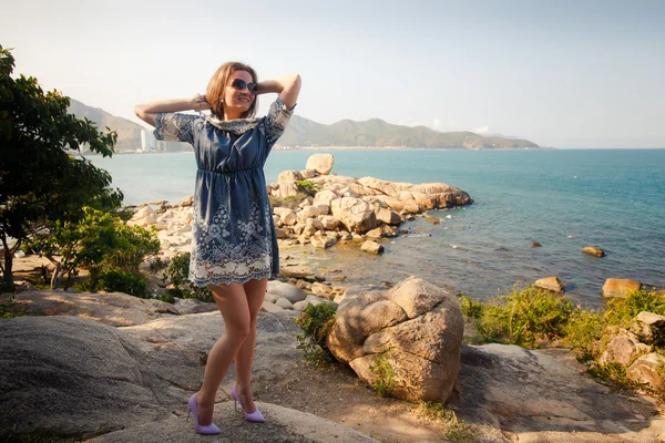 Menina em curto vestido cinza fica em rochas por mar contra a cidade — Fotografia de Stock