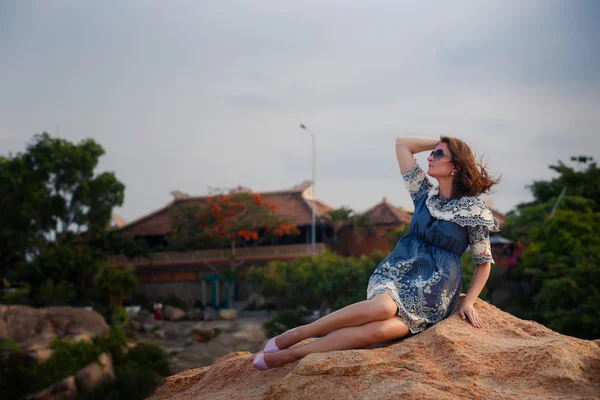 Morena chica en corto gris vestido se sienta en la roca contra villas —  Fotos de Stock