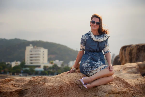 Girl in short grey frock sits on rock shows legs against city — Stock fotografie