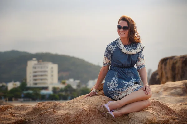 Girl in short grey frock sits on rock shows legs against city — Stock fotografie