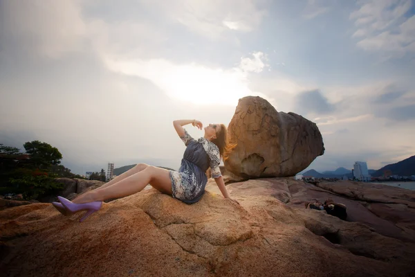 Brunette girl in short grey frock sits on rock against sky — Zdjęcie stockowe