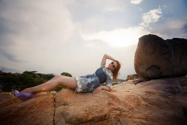 Morena chica en corto gris vestido se sienta en la roca contra el cielo —  Fotos de Stock