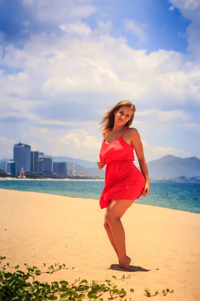 Blond girl in red stands barefoot on sand touches hip under wind — Stock Photo, Image