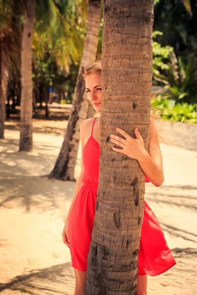 Blond girl in red looks out of palm looks forward against plants — Stock Photo, Image